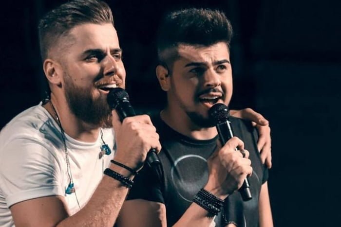 Foto colorida de Zé Neto e Cristiano em fundo preto enquanto cantam no microfone - Metrópoles