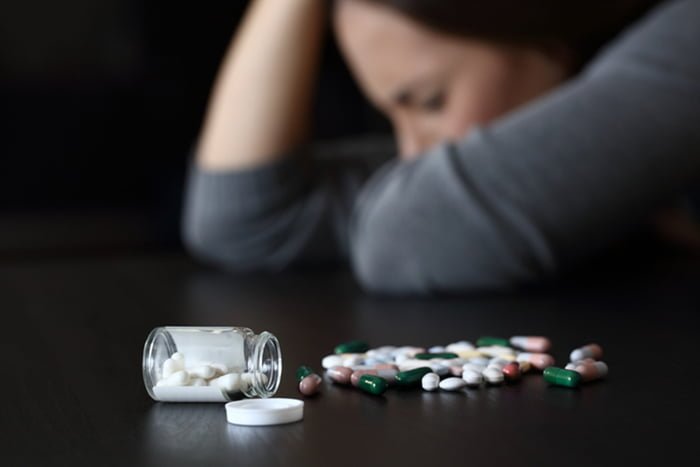 Imagem colorida: remédios antidepressivos sobre a mesa e mulher no fundo com a mão na cabeça - Metrópoles
