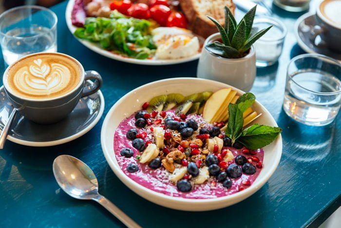 Mesa de café da manhã com xícara de capuccino, frutas, água e talheres