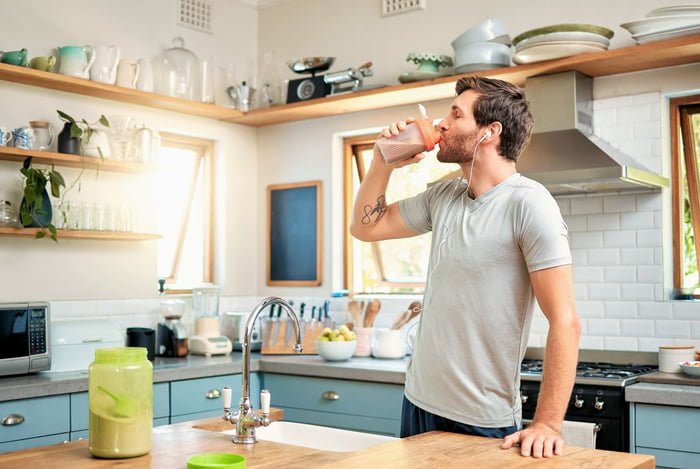Imagem colorida de jovem tomando whey protein na cozinha de uma casa - Metrópoles