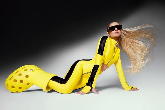 Em fundo neutro, Paris Hilton apoiada no chão. Ela usa macacão amarelo com detalhes em preto e bota amarela gigante - Metrópoles