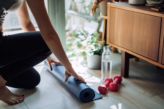 Foto mostra mulher enrolando tapete de yoga em casa ao lado de pesos