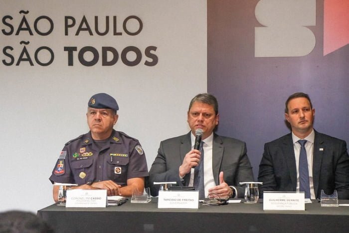 Foto colorida do governador Tarcísio de Freitas (de terno cinza), entre o coronel Cássio (à esquerda, de farda), e o secretário Guilherme Derrite (à direita, de terno preto)
