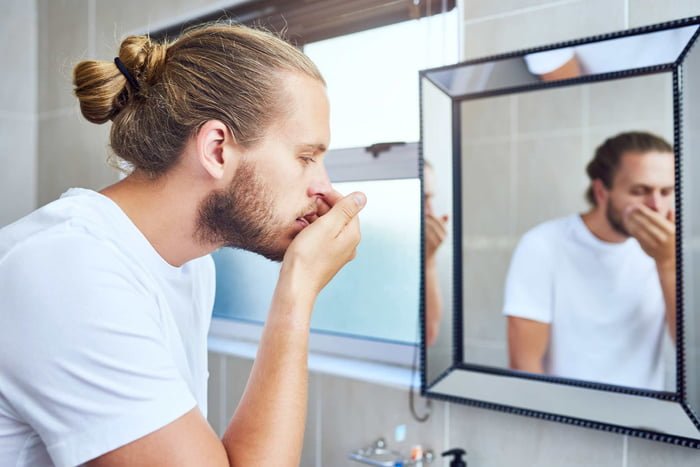 Foto mostra homem loiro diante do espelho verificando a respiração para saber se está com mau hálito