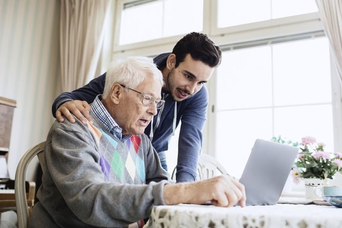 Foto colorida de idoso pardo mexendo em um notebook, colocando em cima de uma mesa. Ao lado do idoso, está um homem jovem e branco. Ele está com a mão no ombro do idoso - Metrópoles