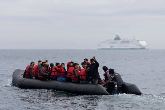 imagem colorida mostra barco em alto mar cheio de imigrantes com colete salva vidas, com navio ao fundo - Metrópoles