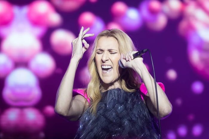 Foto colorida da cantora Céline Dion cantando com um micrfone na mão e os olhos fechados em um fundo de led colorido - Metrópoles