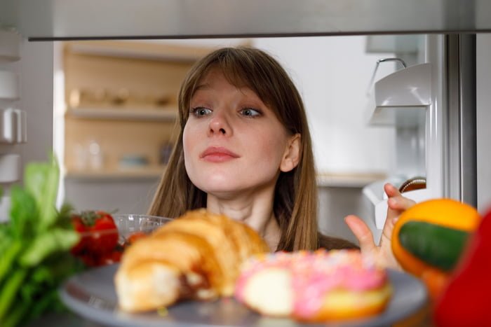 Foto colorida de mulher branca olhando alimentos dentro da geladeira - Metrópoles