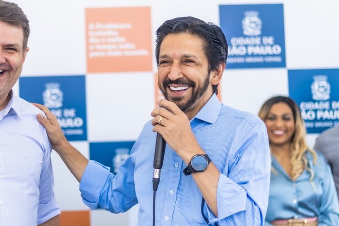 Imagem colorida mostra Ricardo Nunes sorrindo, falando ao microfone, com a mão direita no ombro de um aliado, na frente de um palco com um banner da Prefeitura de São Paulo. Nunes é branco, de meia idade, tem cabelo e barba pretos e veste uma camisa azul, com as mangas arregaçadas - Metrópoles