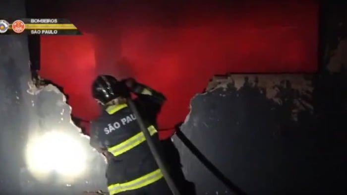 Imagem de bombeiro segurando mangueira em direção a chamas dentro de um prédio - Metrópoles
