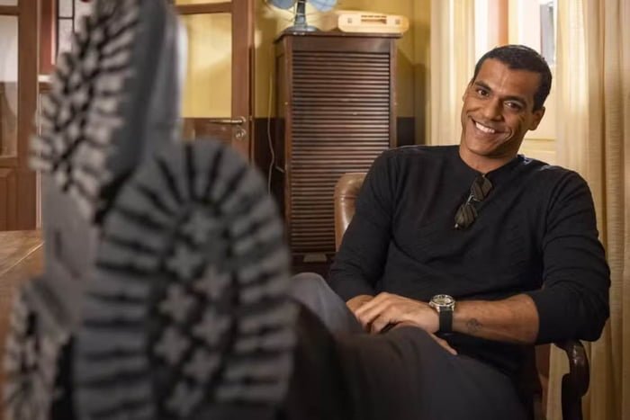 Foto de um homem negro, usando roupa toda preta, e sorrindo. Ele está sentado em uma cadeira - Metrópoles