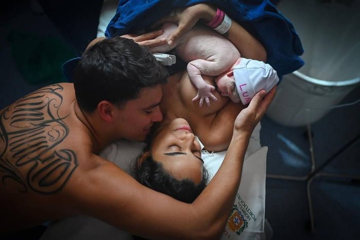foto colorida de mulher deitada com bebe no peito e homem acariciando o bebe - metrópoles