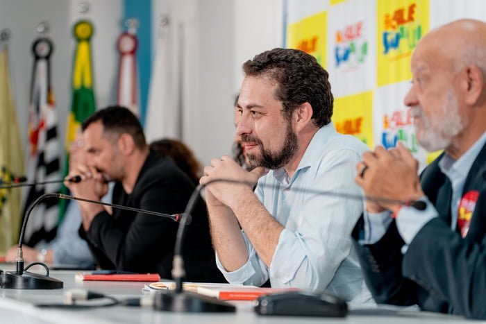 Imagem colorida mostra Boulos sentado em uma mesa de reuniões, com as mãos no queixo, sorrindo - Metrópoles
