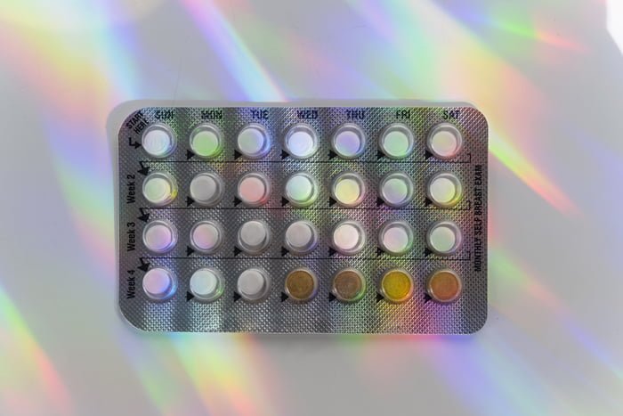 Fotografia mostra cartela de anticoncepcional em fundo branco, com luzes coloridas - Metrópoles