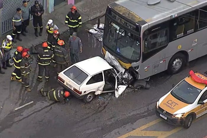 Imagem de aérea de carro branco destruído após bater em ônibus; oito agentes inspecionam o local - Metrópoles