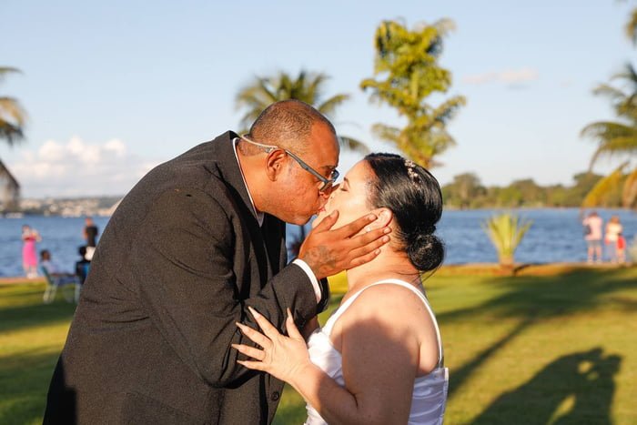 Foto colorida de casal, homem e mulher, de terno e vestido de noiva, se beijando às margens de lago
