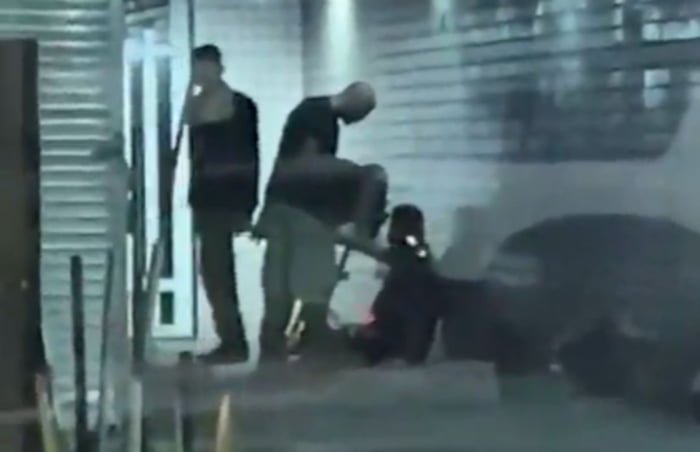 Imagem colorida mostra, de longe, homem caído no chão enquanto homens em pé apontam dedo para a cara dele