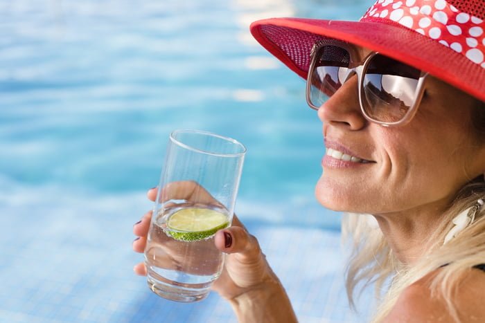 Foto de uma mulher sorridente segurando um copo de água com limão na beira de uma piscina - Metrópoles