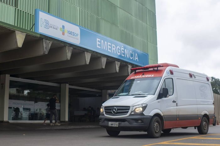 Fachada do Hospital de Base do Distrito Federal, com ambulãncia na frente de porta da emergência
