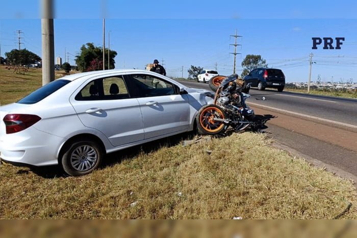 Imagem colorida de um carro branco no canteiro de uma rodovia, com uma moto caída em frente ao veículo após acidente entre os dois automóveis