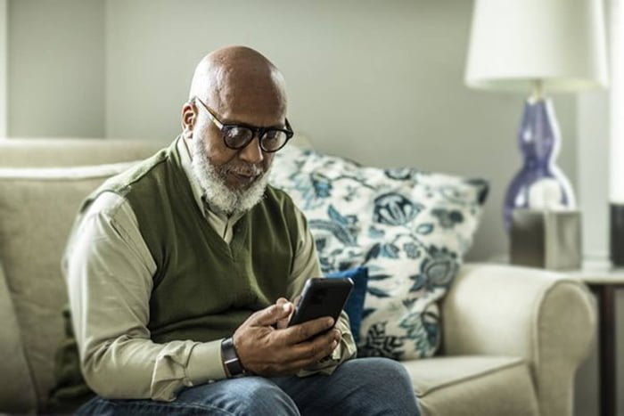 Imagem colorida de um senhor preto sentado no sofá com um celular na mão